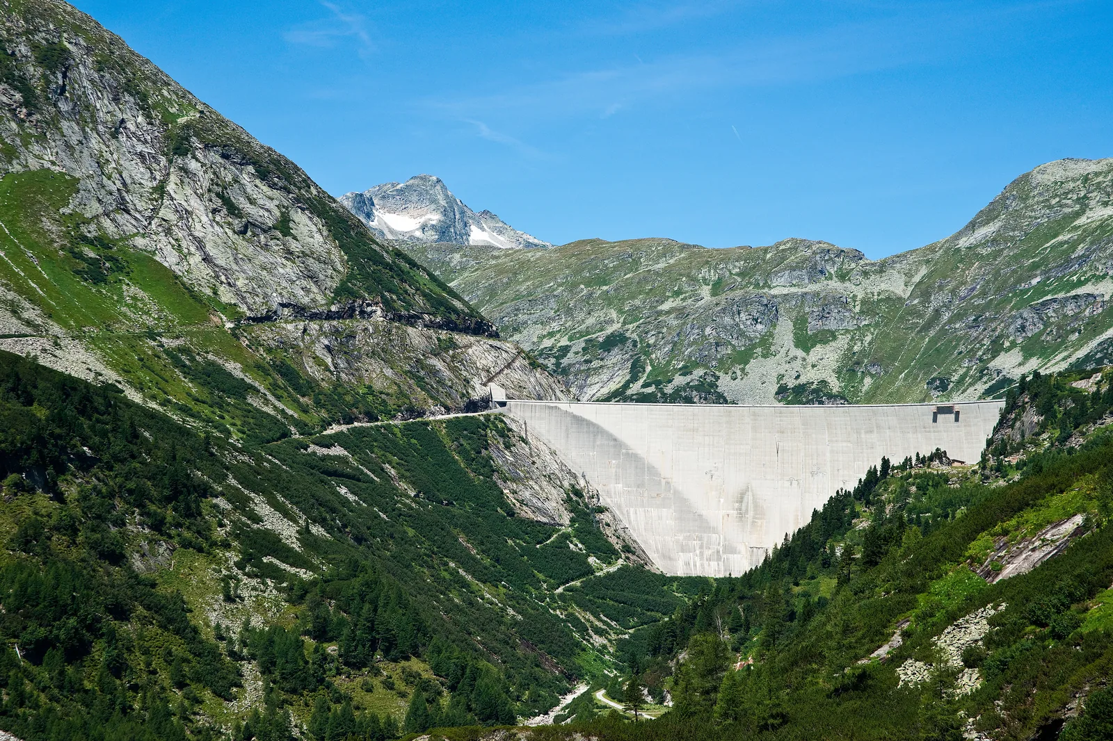 Curved monumental concrete arch dam wall holding back an expansive alpine reservoir among rugged granite mountain peaks.