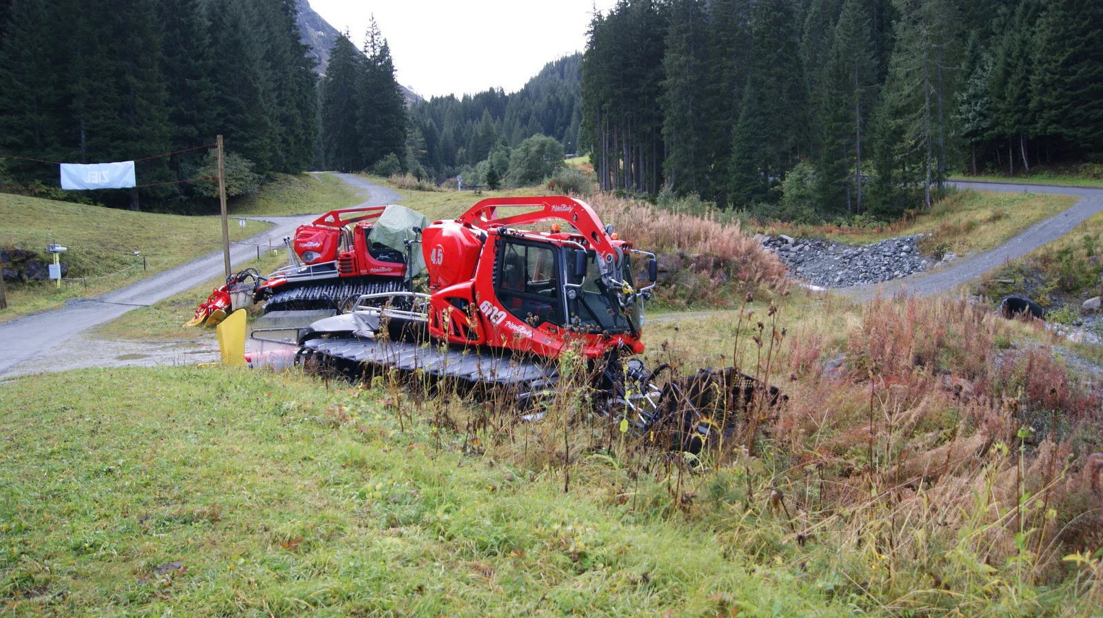 Red caterpillar-tracked snow groomer vehicle resting beside alpine ski slopes in the Austrian mountains.
