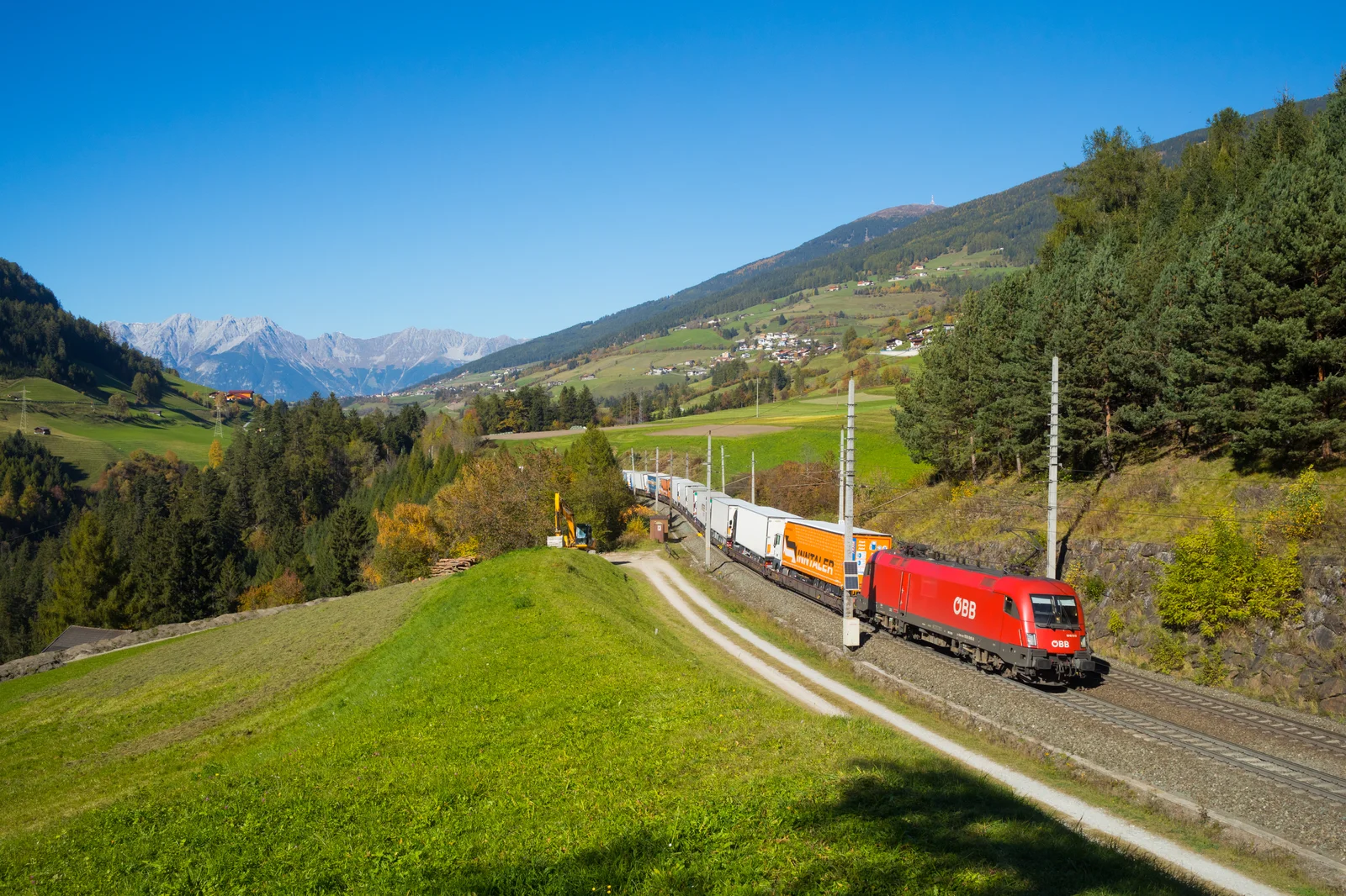 Heavy freight train carrying tractor-trailer trucks on flatcars winding through a scenic alpine valley in Tyrol.