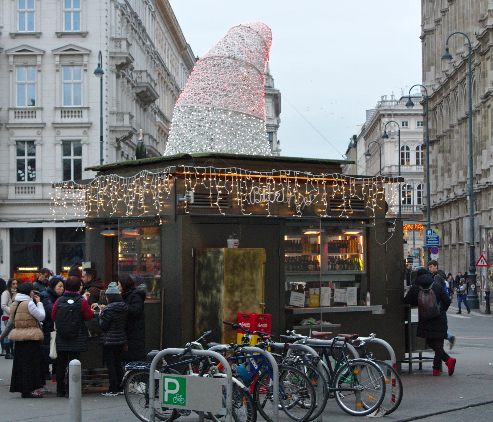 A bustling Viennese sausage stand glowing at night in front of historic architecture.