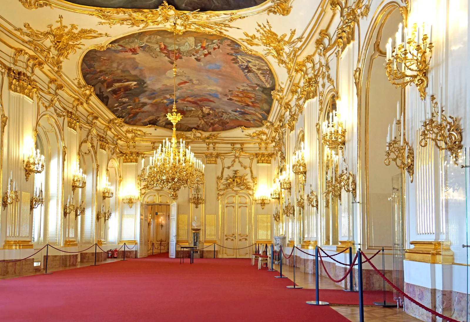 Gilded rococo ballroom with tall arched windows, ornamental white plaster, and elaborate painted ceilings.