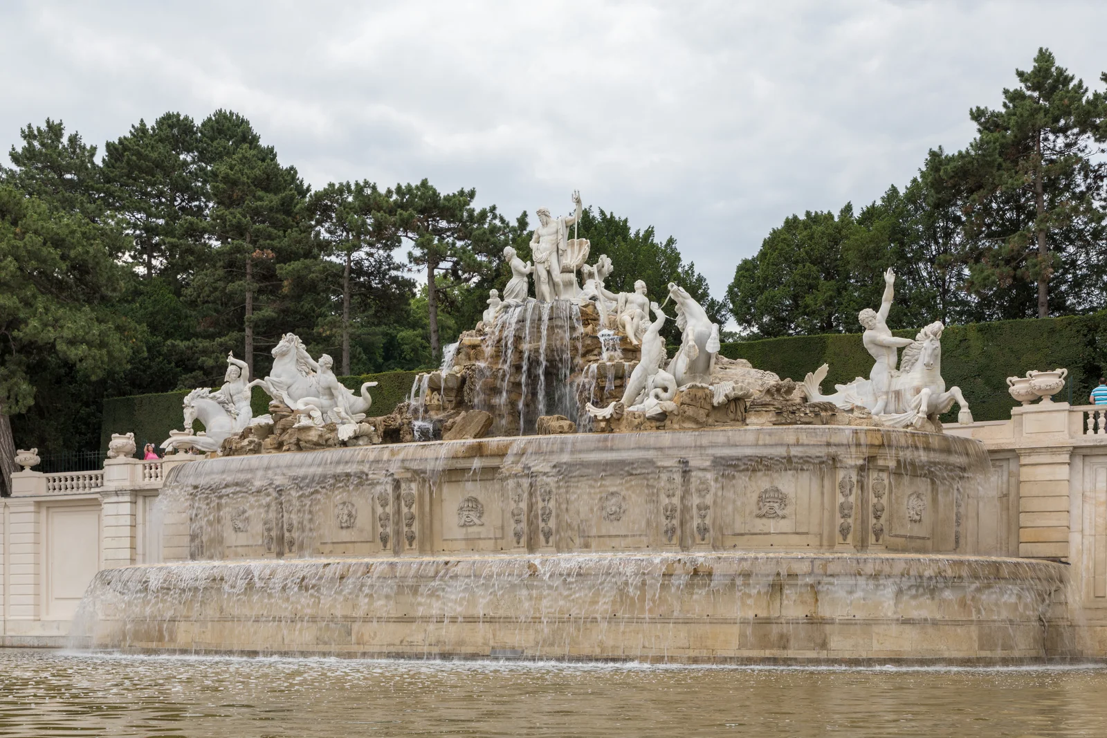 Monumental stone fountain featuring sea deities and horse sculptures beneath a green hillside arcade.