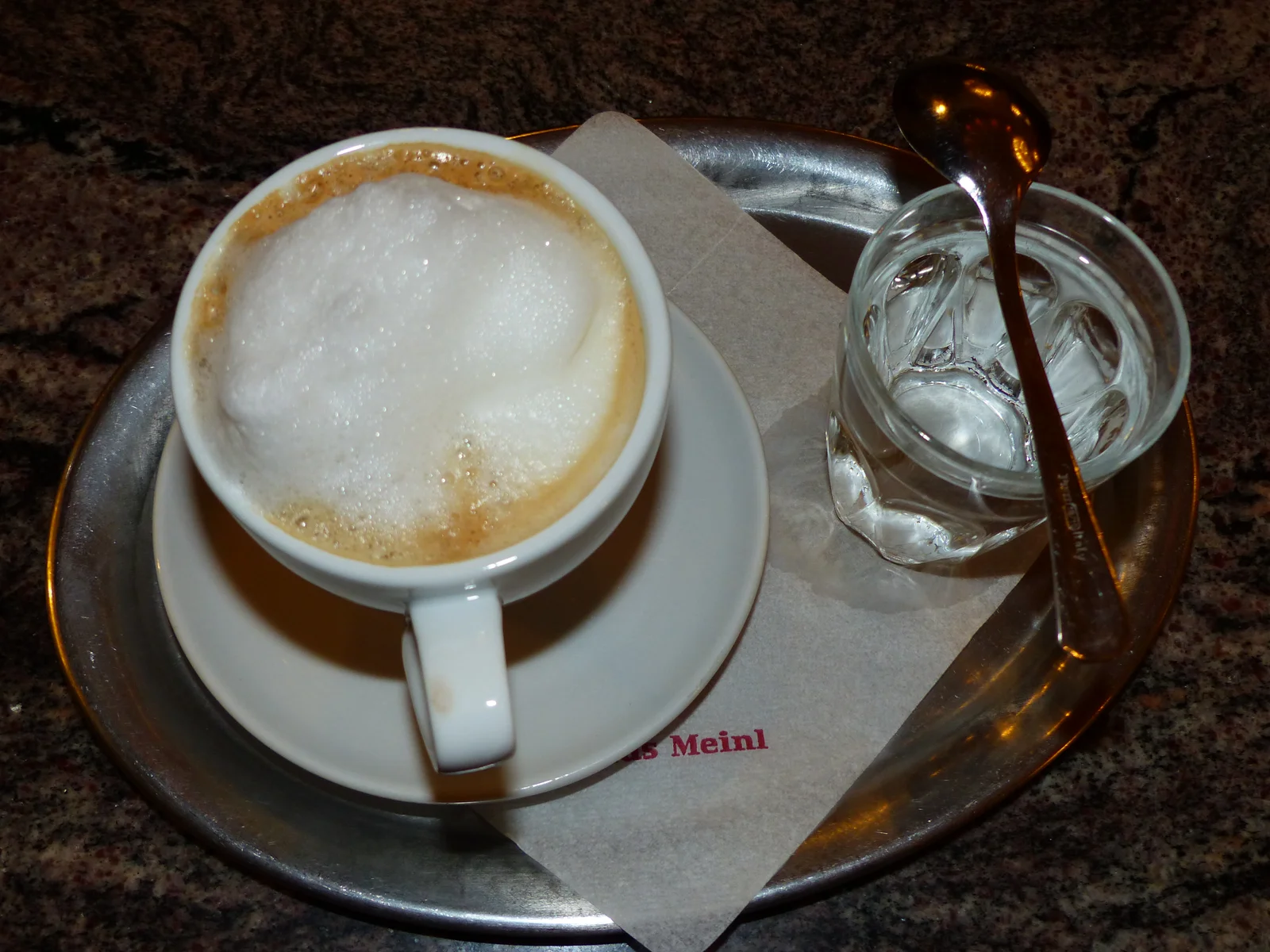 Coffee cup with foamed milk served on a metal tray with a water glass in an Austrian cafe.
