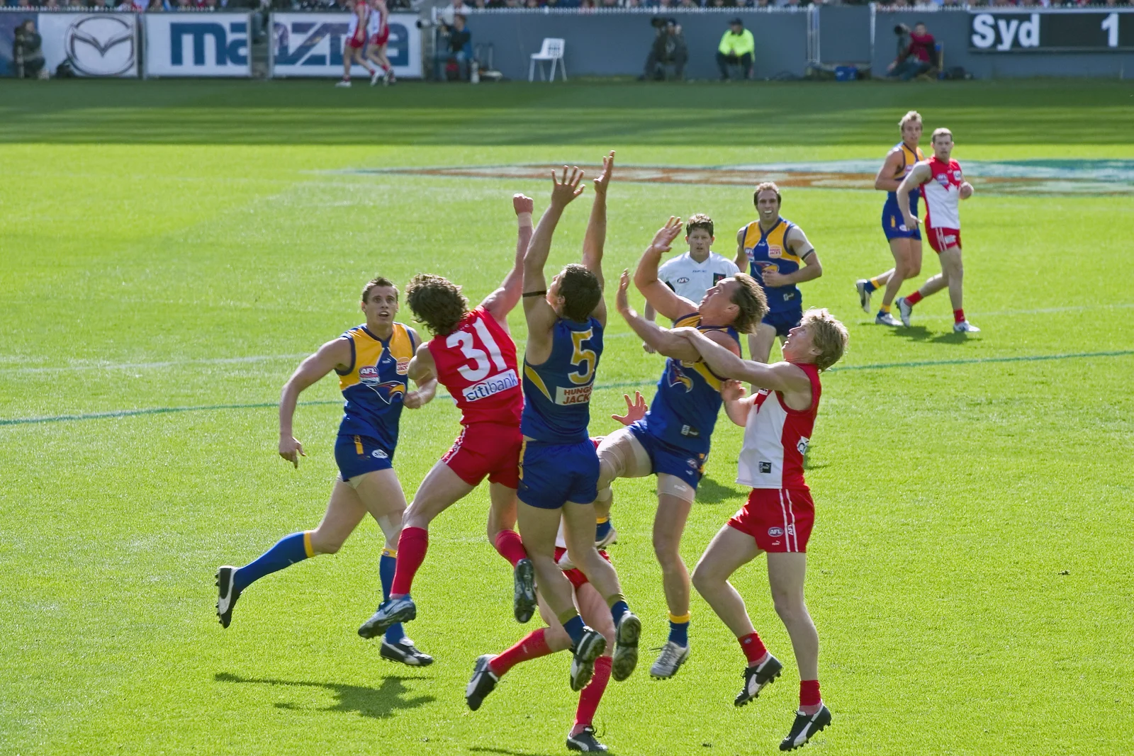 Australian Rules footballers leaping high into the air to catch an aerial mark above opposing players.