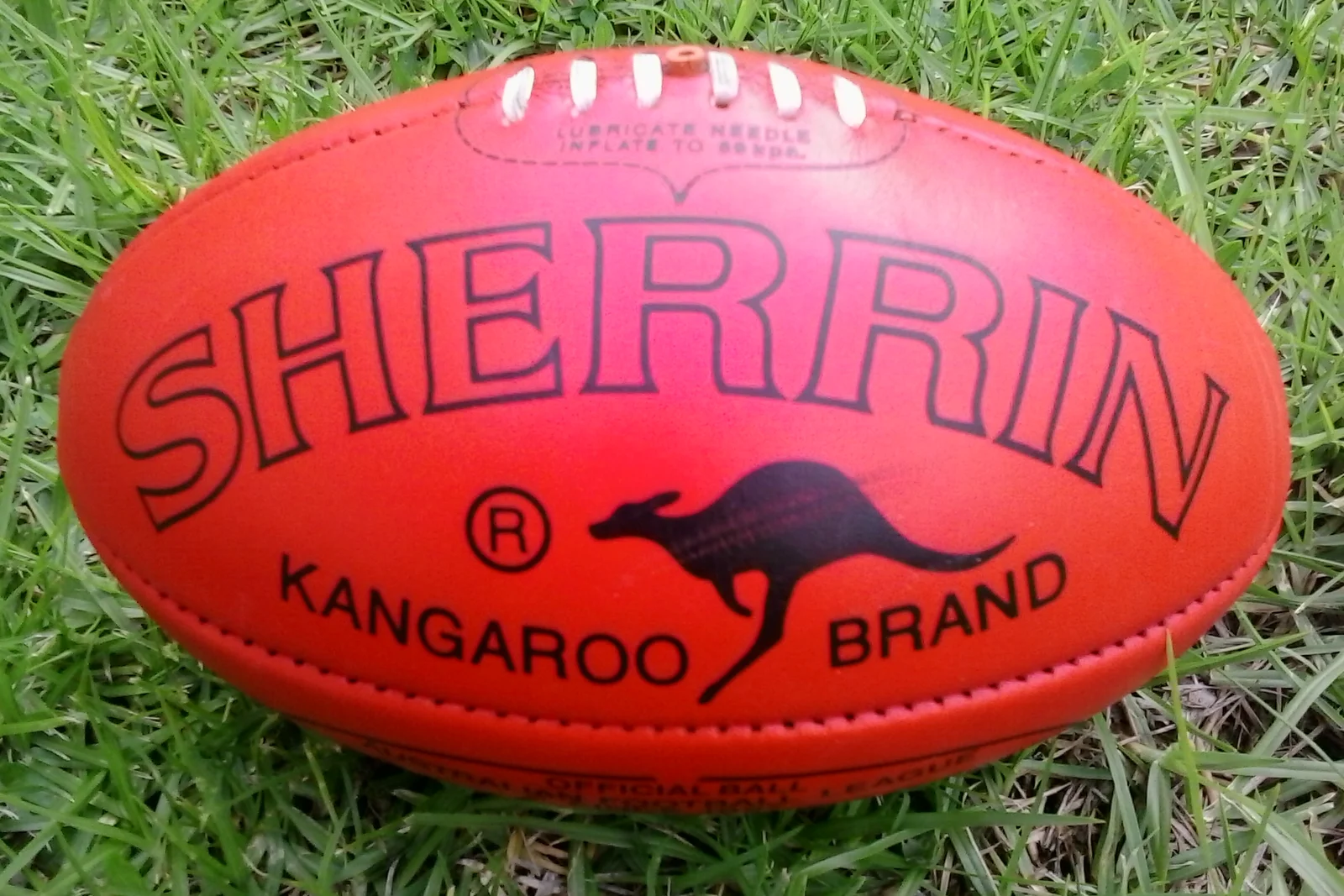 A red leather Sherrin Australian Rules football resting on green grass with visible cross lacing.