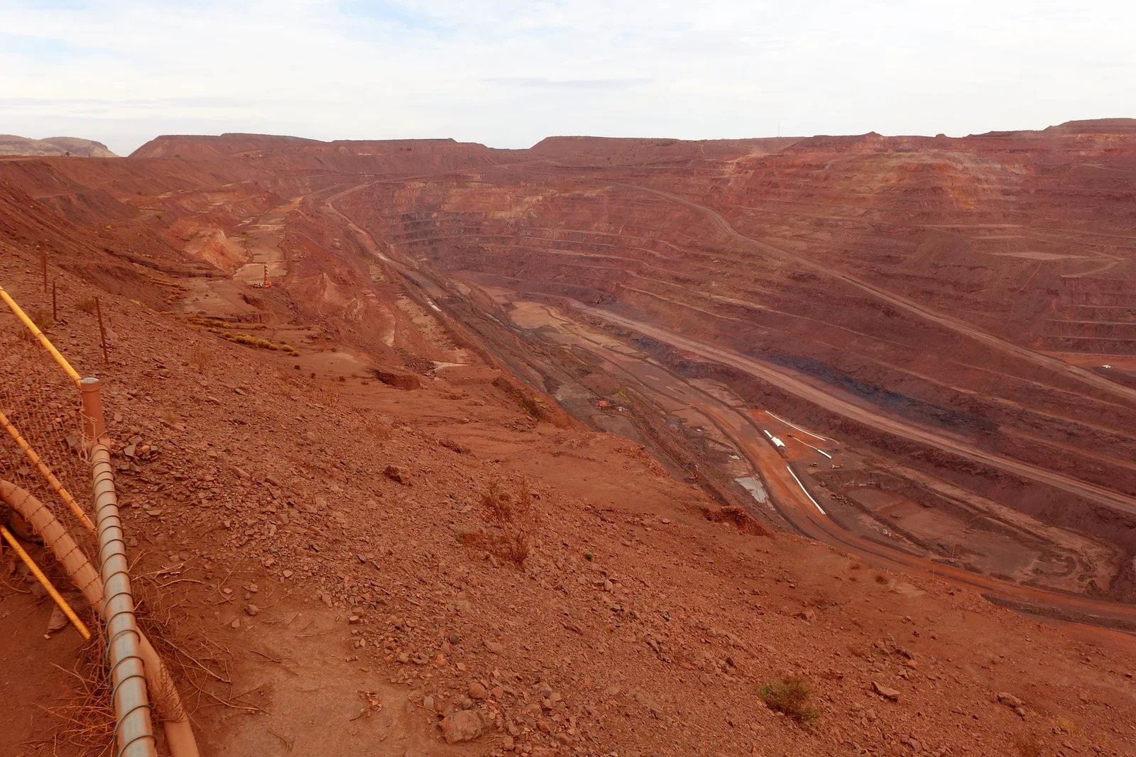 Vast red open-cut iron ore mine with stepped benches and extraction roads under a clear sky.