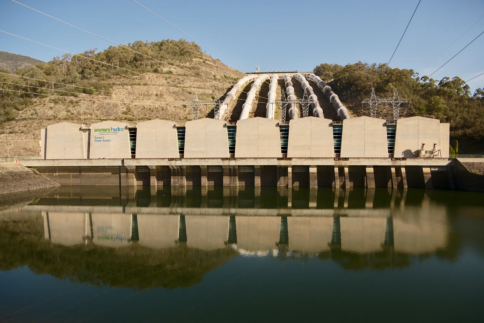 Six massive surface penstock pipes leading down a forested mountain slope into a large hydroelectric power station.