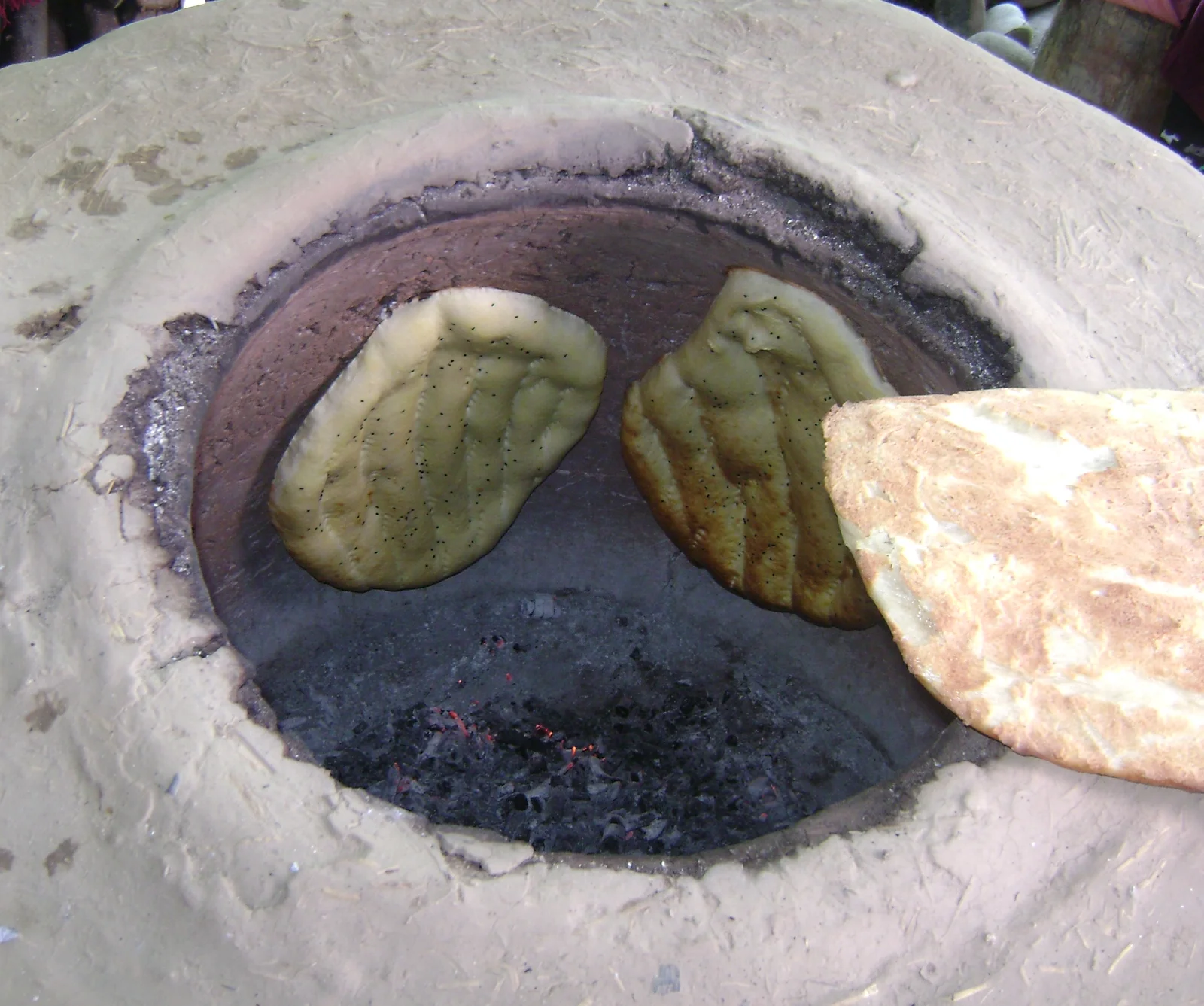 An Azerbaijani woman baker in traditional headscarf bending over a clay tandir oven to slap dough onto the interior wall.