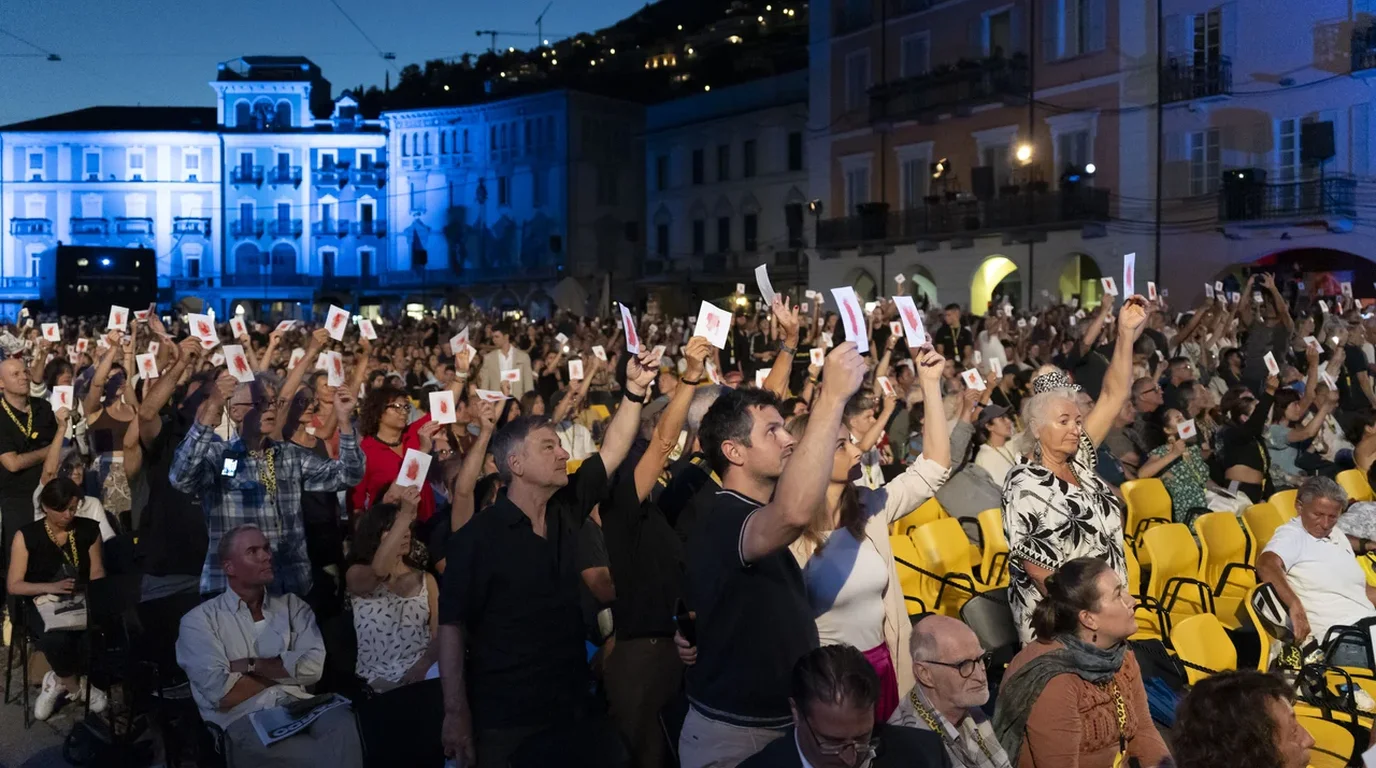 A vast open-air cinema venue filled with thousands of seated spectators facing an illuminated colossal movie projection screen at night.
