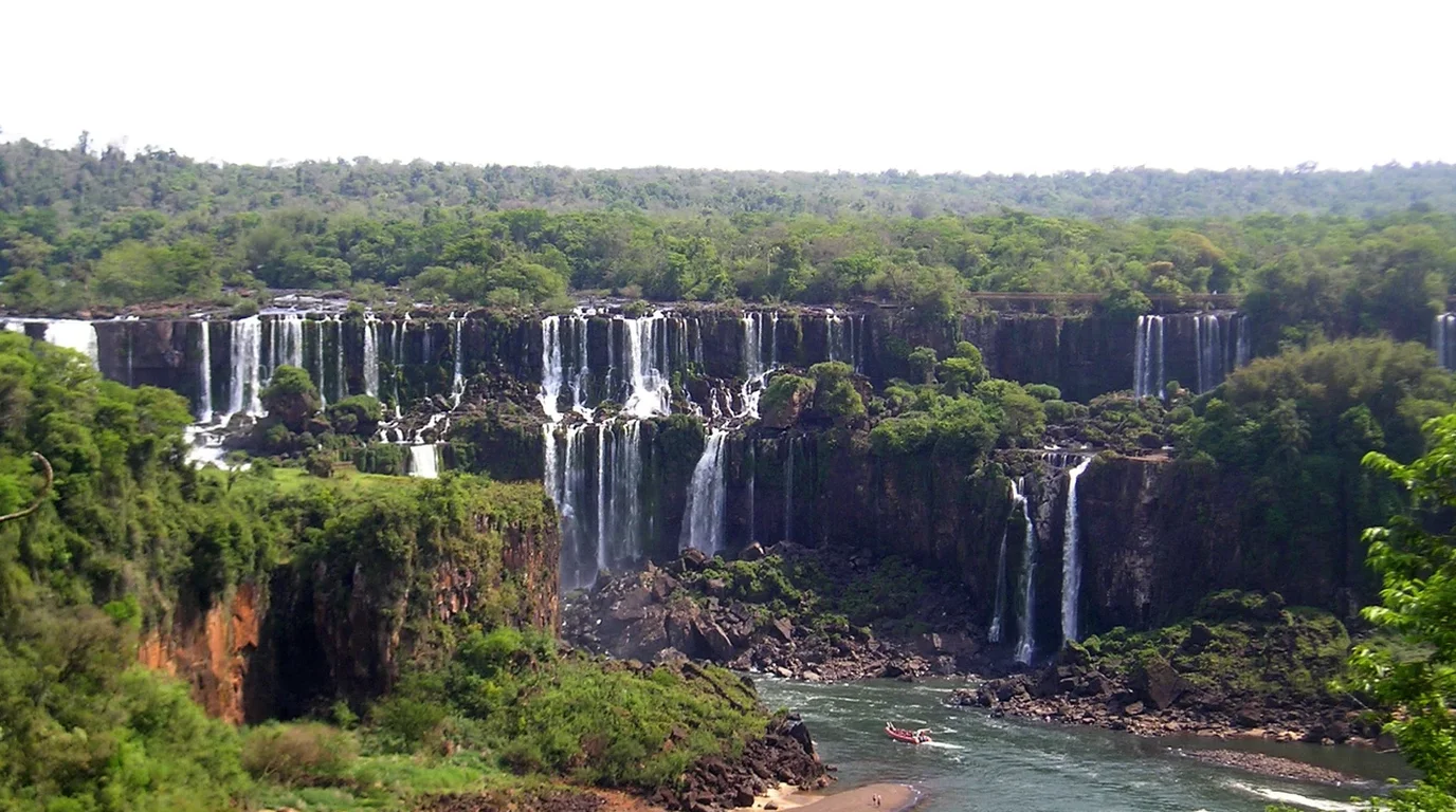 A wide, powerful multi-tiered waterfall cascading over terraced natural limestone barriers surrounded by dense green forest.
