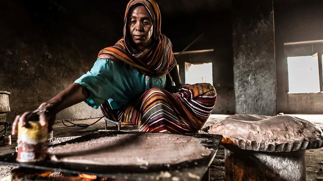 A Sudanese woman tending the charcoal embers of a traditional baking hearth while preparing flatbread.