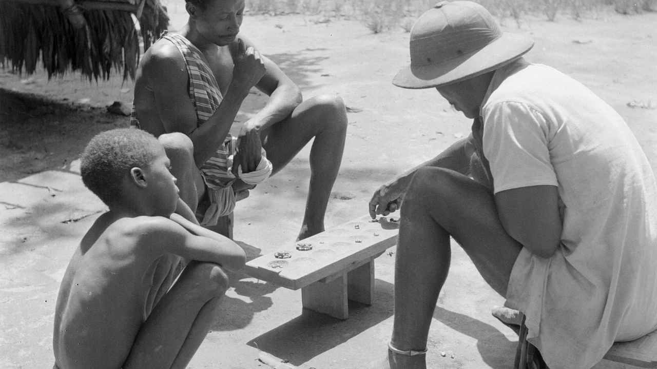Two players seated outdoors across a carved wooden board playing the traditional Ba game of strategy in Suriname while a boy observes.