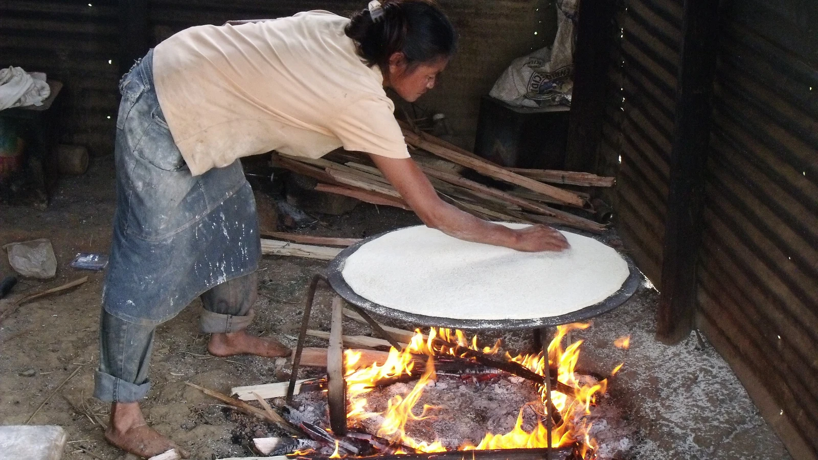 An indigenous baker baking circular flat cassava bread over an open-fire ceramic griddle.