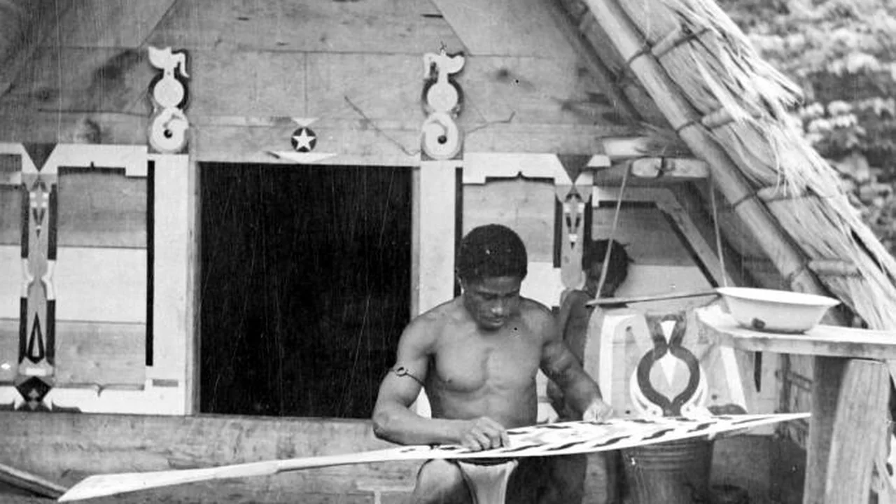 A Maroon woodcarver in Suriname meticulously carving and painting an ornate ceremonial paddle in front of a carved timber home.