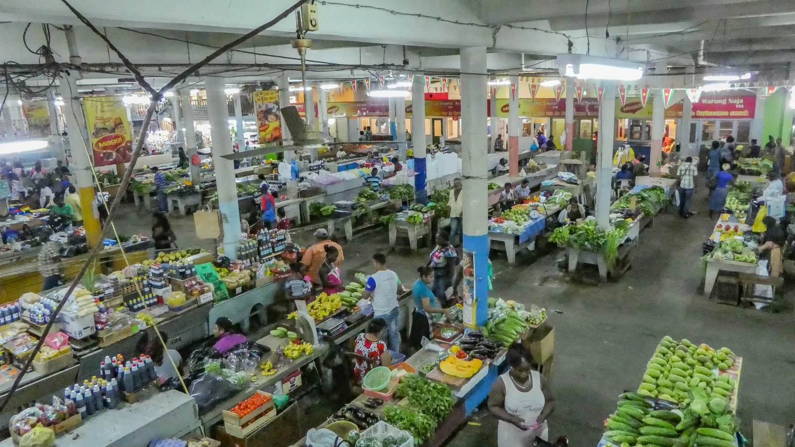 Bustling covered hall of Paramaribo Central Market filled with vegetable stalls and shoppers.