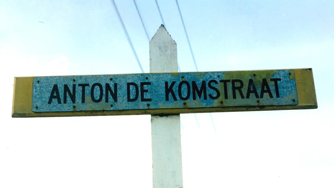 Official blue and white Dutch street name sign for Anton de Komstraat mounted on a timber post in Paramaribo.